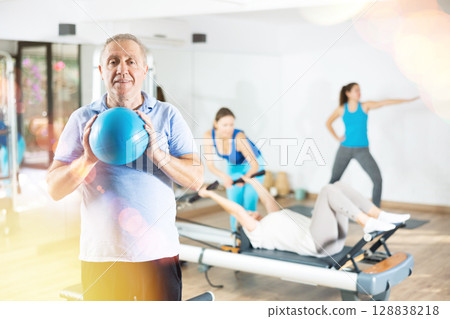 Smiling mature male squeezing ball in hands during Pilates training in gym 128838218