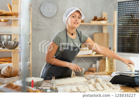 Baker girl cuts small pieces of dough from large lump and puts them on scales. 128838227