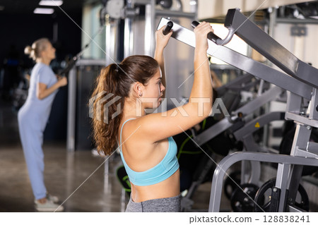 Young female training trains the muscles of the arms and press on simulator in gym. Sport concept Young female training trains the muscles of the arms and press on simulator in gym. Sport concept 128838241