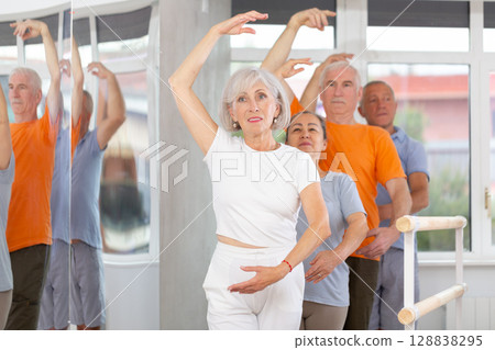 Active mature woman practicing third position holding by ballet barre together with others 128838295
