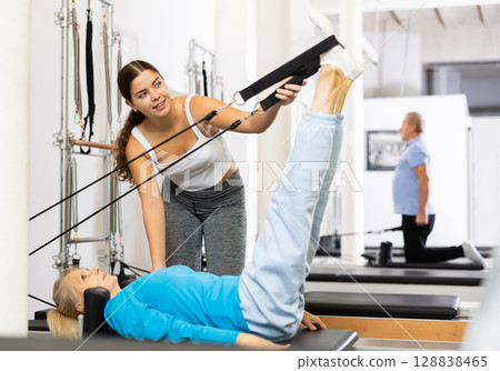Positive young female instructor controlling movements of mature woman exercising on Pilates reformer equipment in studio Positive young female instructor controlling movements of mature woman exercising on Pilates reformer equipment in studio 128838465