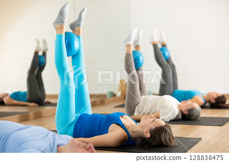 Middle-aged woman practicing pilates with ball in gym area 128838475