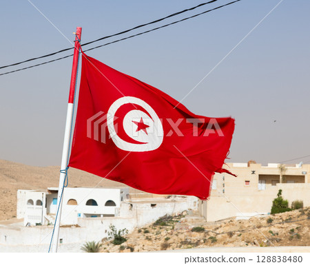 Flag of Tunisia waving against background of stone houses during daytime Flag of Tunisia waving against background of stone houses during daytime 128838480