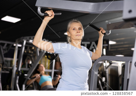 Senior woman doing lat pull-down on lever gym machine 128838567