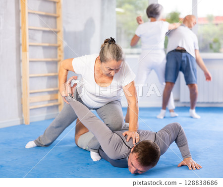 Senior woman in pair with middle-aged coach train Krav Maga and self-defense skills. 128838686