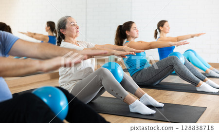 Class of people of different ages holding balls between legs and pushing press during Pilates training in studio 128838718