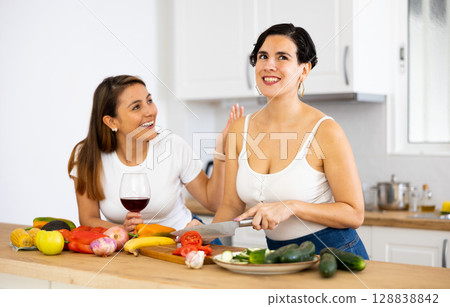 Two happy female friends preparing salad and drinking red wine together in kitchen 128838842