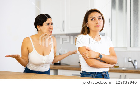Frustrated young woman listening to angry girlfriend in home kitchen Frustrated young woman listening to angry girlfriend in home kitchen 128838940