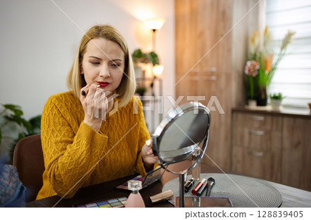 Young woman applying lipstick while seated at a stylish vanity in a cozy modern room with warm lighting 128839405