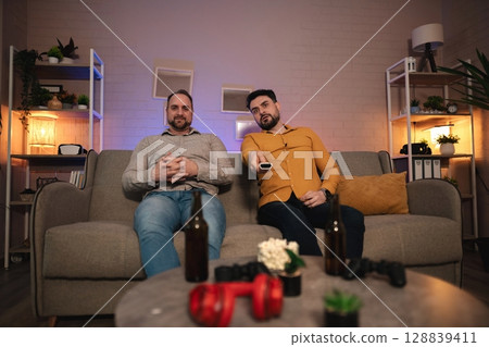 Two friends enjoying a gaming night together in a cozy, well-lit living room with drinks and controllers at hand 128839411