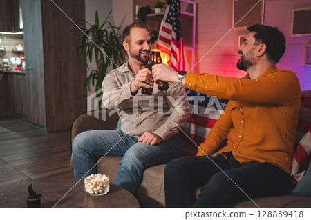 Friends enjoy a lively evening celebration with drinks and laughter in a cozy living room adorned with patriotic decor 128839418