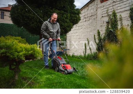 Man mowing a lush green lawn in a serene backyard garden during a cloudy afternoon 128839420
