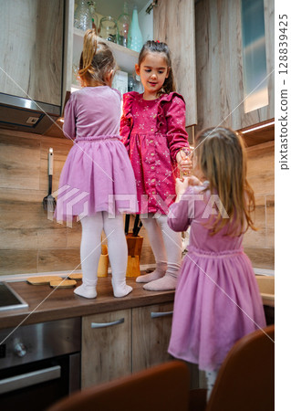 Three young girls in pink dresses explore their kitchen during a playful afternoon gathering full of laughter and discovery 128839425
