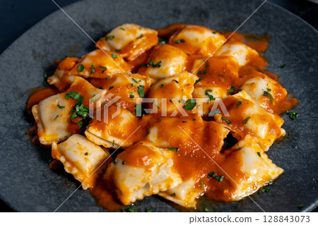 Ravioli pasta with tomato sauce and parsley topping on elegant dark grey plate 128843073