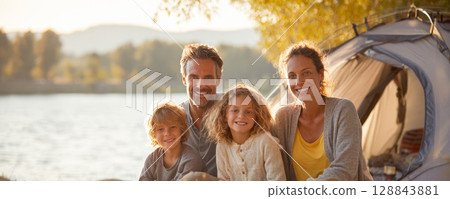 A happy family of four, including two adults, two children, sits together smiling in front of camping tent by tranquil lake at sunset. Warm, golden light suggests peaceful, joyful outdoor adventure. 128843881