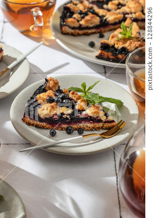 A slice of homemade blueberry crumble pie on a plate on white tile background. Sweet breakfast composition in bright morning light A slice of homemade blueberry crumble pie on a plate on white tile background. Sweet breakfast composition in bright morning light 128844296
