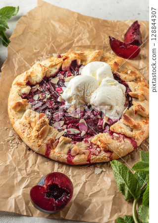 French Galette or pie with plums and almonds served with ice cream scoops on parchment paper background. Summer traditional homemade sweet pastries 128844307
