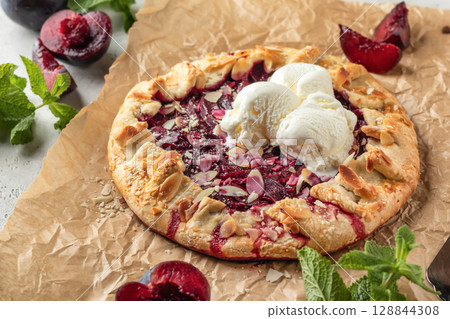 French Galette or pie with plums and almonds served with ice cream scoops on parchment paper background. Summer traditional homemade sweet pastries 128844308