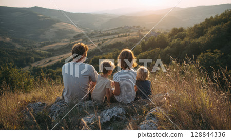A family of four, seen from behind, sits together on a rocky outcrop, admiring a vast mountain landscape bathed in the warm light of a sunset 128844336