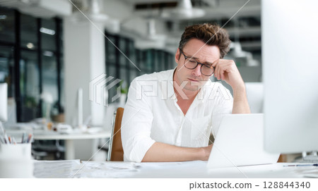 Man wearing glasses, white shirt sits at desk in modern office, looking tired and rubbing his forehead while looking at laptop. Themes of stress, fatigue, challenges of demanding work environment. 128844340