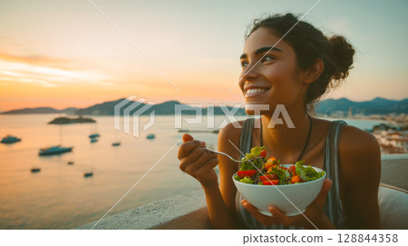 A radiant woman smiles while savoring a vibrant salad, basking in the warm glow of a breathtaking sunset over the sea. Epitomizing healthy living and serene relaxation 128844358