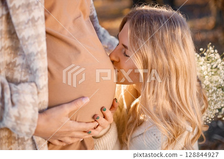 Close-up view of happy girl near a pregnant belly. Women expecting a baby and standing in a autumn park. Portrait of curly blonde woman. Close-up view of happy girl near a pregnant belly. Women expecting a baby and standing in a autumn park. Portrait of curly blonde woman. 128844927