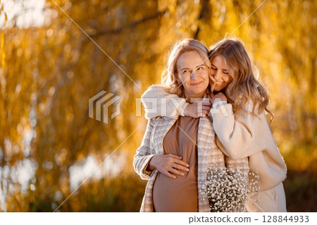 Adult daughter hugs her pregnant mom on a sunny autumn day in nature. Blonde woman holding flowers in her hand. Women wearing beige clothes. 128844933