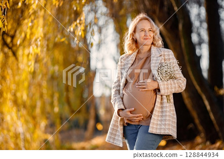 Portrait of middle-aged pregnant woman outdoors at park. Middle age pregnant woman expecting baby at aged pregnancy. Blonde woman wearing brown sweater and beige coat. 128844934