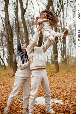 Man, woman and their little girl standing in autumn forest and laughing. Brunette man throwing his daughter in the air. Family wearing beige sportive costumes. 128845047
