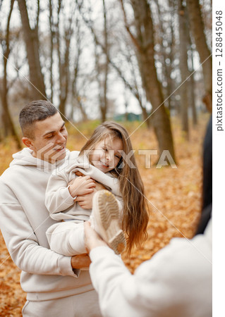 Cropped photo of a man and a little girl playing in autumn forest and laughing. Brunette woman is blurred and man holding their daughter on hands. Family wearing beige sportive costumes. Cropped photo of a man and a little girl playing in autumn forest and laughing. Brunette woman is blurred and man holding their daughter on hands. Family wearing beige sportive costumes. 128845048