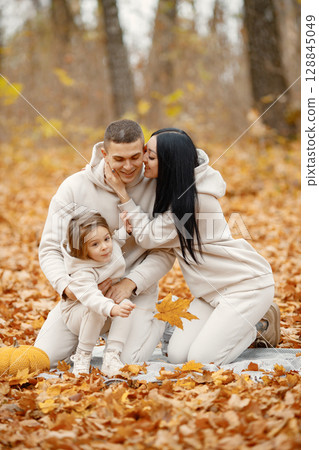 Man, woman and their little girl sitting on a blanket in autumn forest. Brunette woman and man play with daughter. Family wearing beige sportive costumes. 128845049