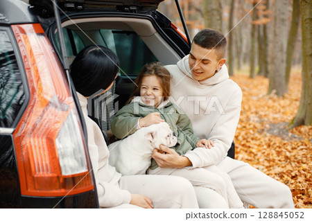 Family resting after day spending outdoor in autumn park. Father, mother, their daughter and dog sitting inside car trunk, smiling. Family wearing beige sportive costumes. Family resting after day spending outdoor in autumn park. Father, mother, their daughter and dog sitting inside car trunk, smiling. Family wearing beige sportive costumes. 128845052