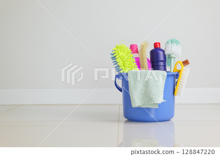 Cleaning supplies in a blue bucket on the floor. Essential cleaning tools like detergent bottles, brushes, and sponges are ready for housework in the living room, making home cleaning more efficient. 128847220