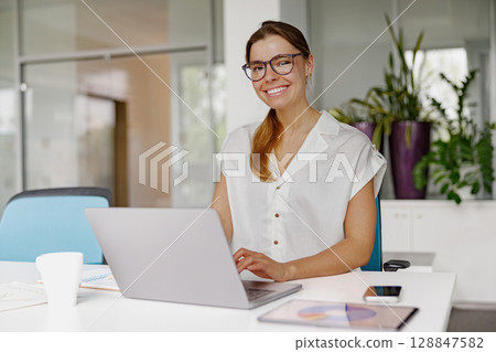 A happy, smiling individual is diligently working on a sleek laptop within a modern office setting 128847582