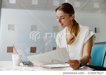 A professional businesswoman is meticulously analyzing various documents in a modern office setting A professional businesswoman is meticulously analyzing various documents in a modern office setting 128847627