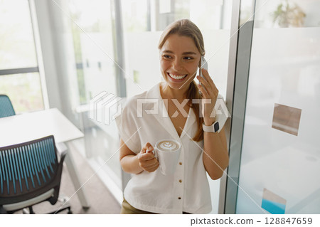 A smiling woman in a modern office is talking on the phone while enjoying her coffee beverage A smiling woman in a modern office is talking on the phone while enjoying her coffee beverage 128847659