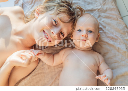 Older and younger baby brothers lying on a soft muslin blanket in warm sunlight, bonding and interacting. Concept of sibling love, early connection, and family warmth 128847935