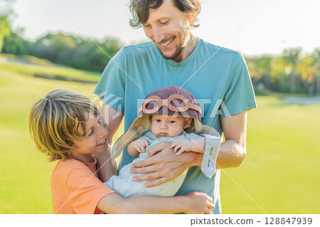 Father, son, and newborn on a meadow. Baby wearing a pilot hat, symbolizing dreams of aviation. Concept of family bonding, future pilot, and childhood aspirations Father, son, and newborn on a meadow. Baby wearing a pilot hat, symbolizing dreams of aviation. Concept of family bonding, future pilot, and childhood aspirations 128847939