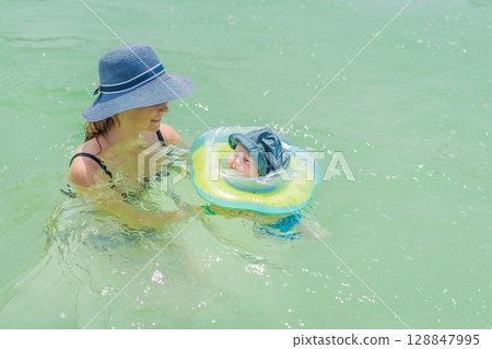 Baby floating in a neck ring in turquoise water with mom nearby. Early swimming and water safety concept 128847995