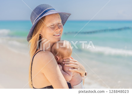 Mother holding her newborn baby by the turquoise sea, sharing a peaceful and loving moment. Family bonding, motherhood, and travel with baby concept 128848023