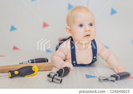 Adorable baby in a mechanic costume surrounded by toy tools. Concept of repair, imaginative play, and introducing kids to the world of fixing and problem-solving through fun 128848025