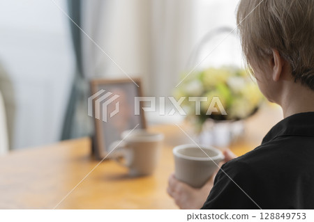 A woman having tea time in the living room while looking at a portrait of her husband 128849753