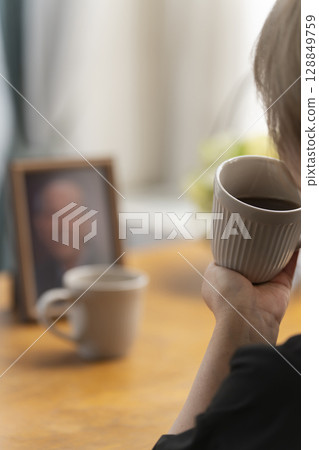 A woman having tea time in the living room while looking at a portrait of her husband A woman having tea time in the living room while looking at a portrait of her husband 128849759