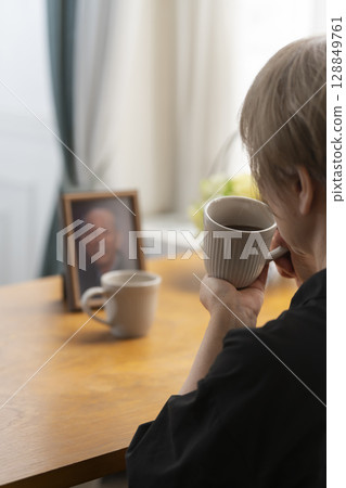 A woman having tea time in the living room while looking at a portrait of her husband A woman having tea time in the living room while looking at a portrait of her husband 128849761