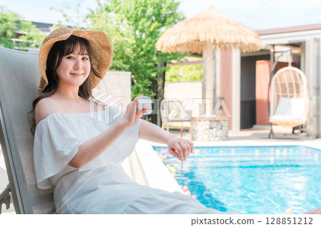 一位年輕的亞洲女性坐在泳池邊的躺椅上，在手臂上塗抹防曬霜 128851212