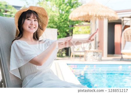 一位年輕的亞洲女性坐在泳池邊的躺椅上,在手臂上塗抹防曬霜 一位年輕的亞洲女性坐在泳池邊的躺椅上,在手臂上塗抹防曬霜 128851217