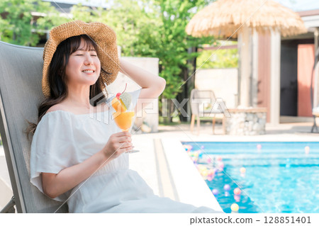 Young Asian woman sitting on a recliner by the pool and drinking tropical juice Young Asian woman sitting on a recliner by the pool and drinking tropical juice 128851401