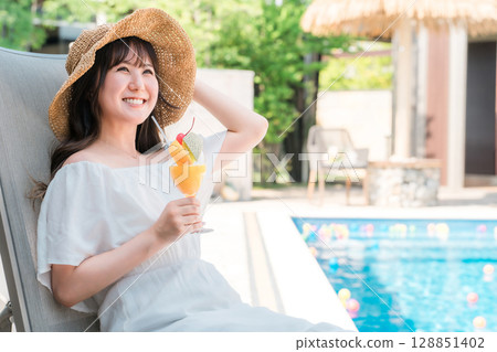 Young Asian woman sitting on a recliner by the pool and drinking tropical juice Young Asian woman sitting on a recliner by the pool and drinking tropical juice 128851402