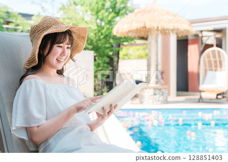 一位年輕的亞洲女性坐在泳池邊的躺椅上看書 128851403