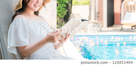一位年輕的亞洲女性坐在泳池邊的躺椅上看書 128851404